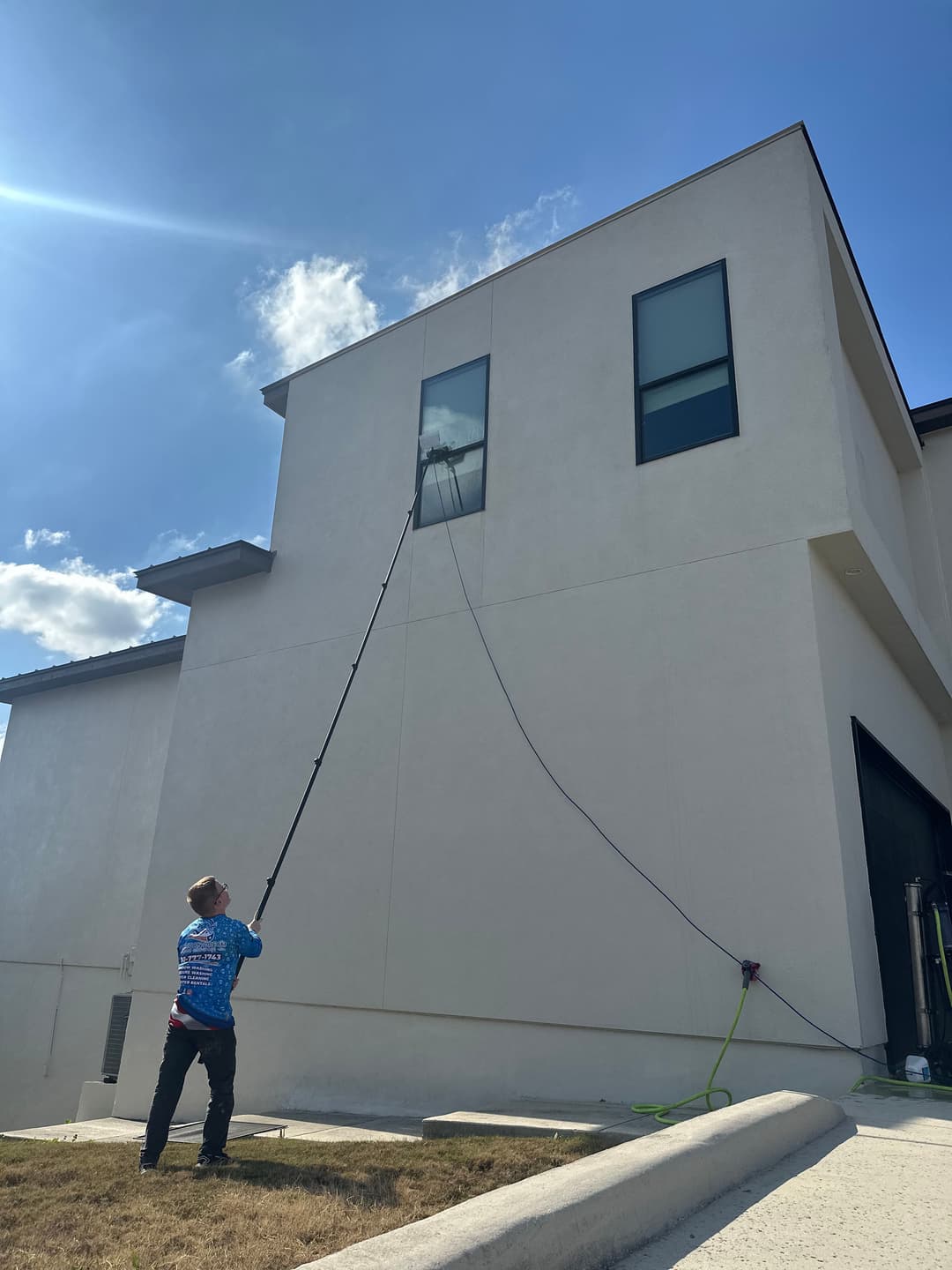 Man using a pole to clean a high window on a modern building under a clear blue sky.