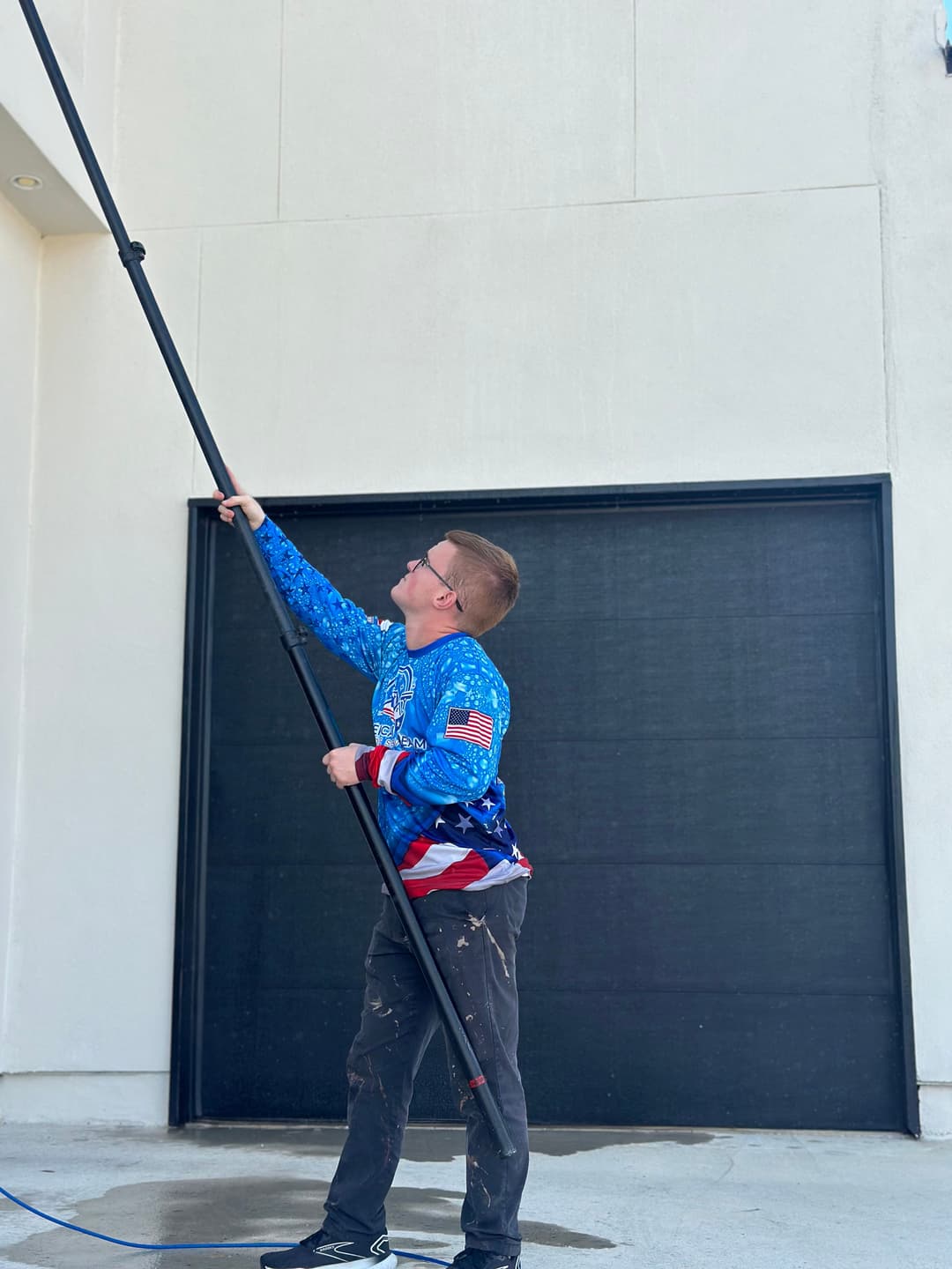 Man painting exterior wall with extension pole, wearing a blue shirt with American flag design.