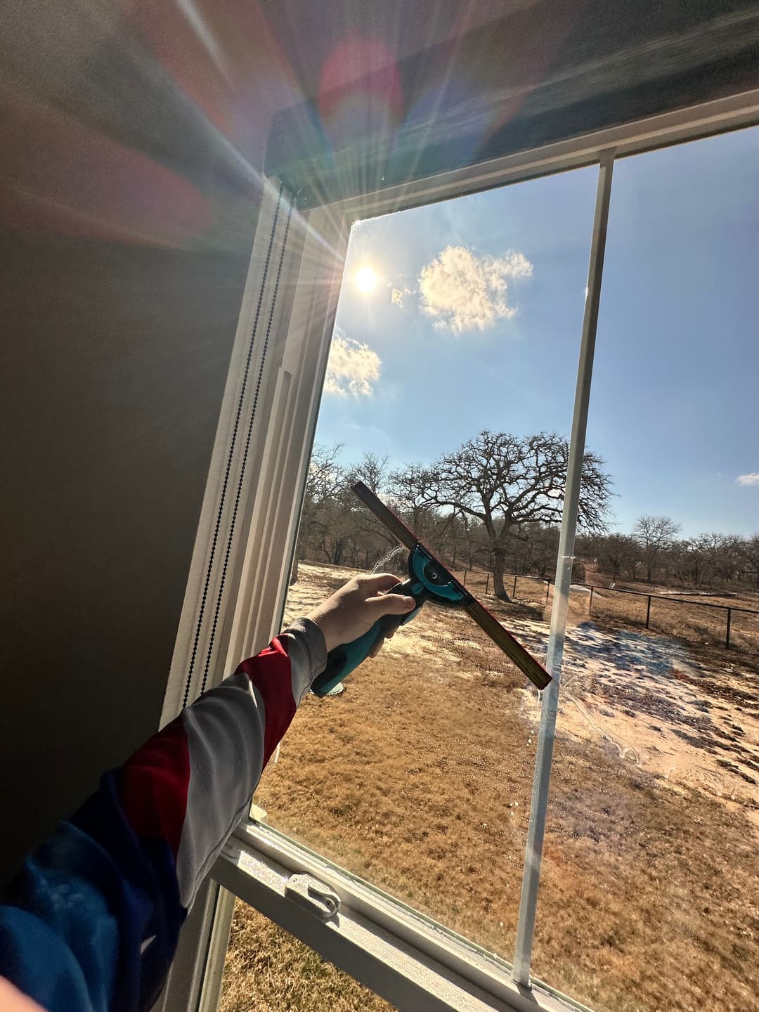 Person cleaning a window with a squeegee, bright sun and landscape visible outside.