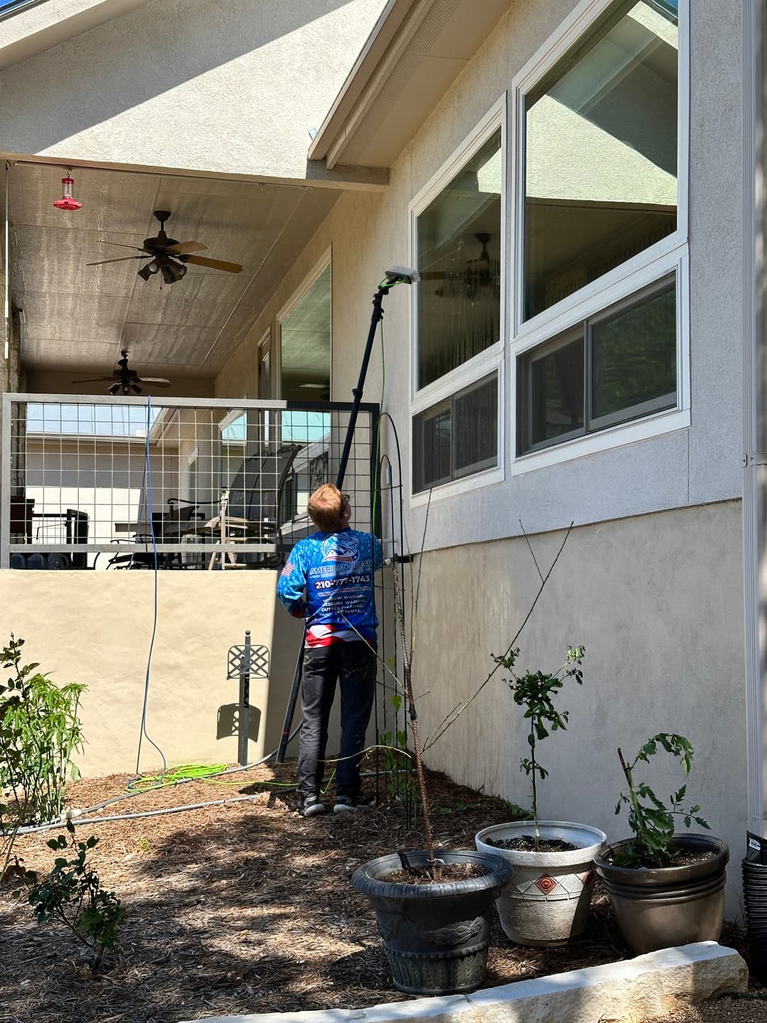 Person using a pole to clean windows on a house, with garden plants and patio visible.