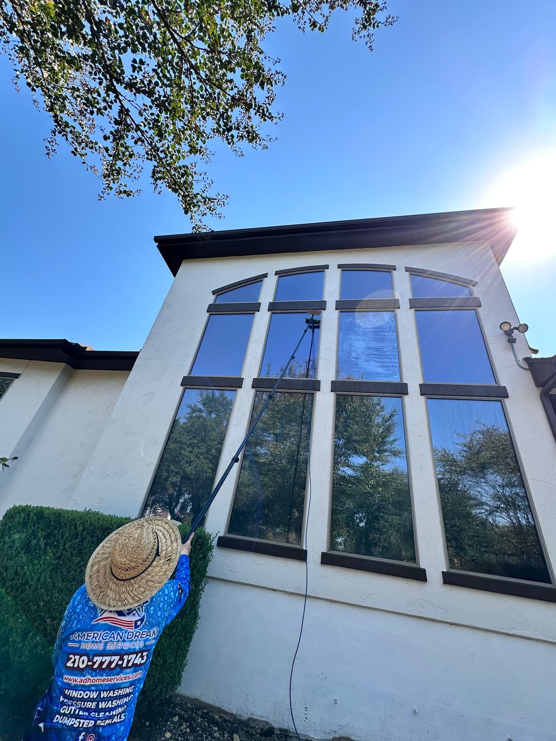 Professional window cleaner using a telescopic pole on large windows of a house under blue sky.