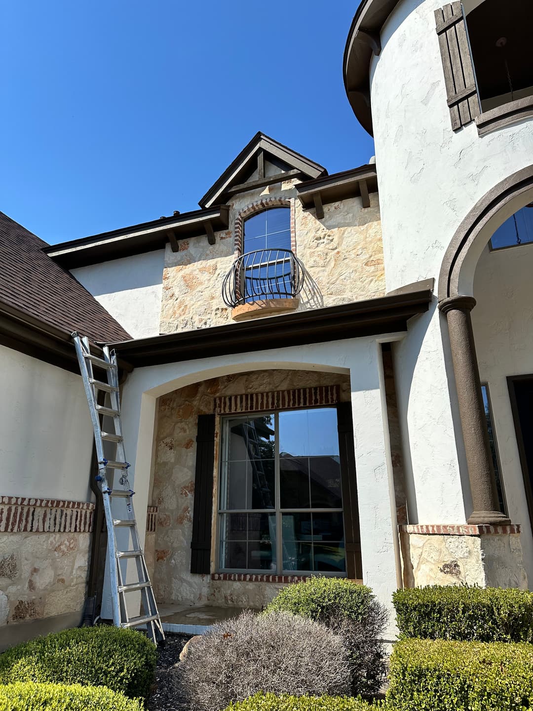 Ladder positioned beside a house with stone facade and decorative window balcony under clear blue sky.