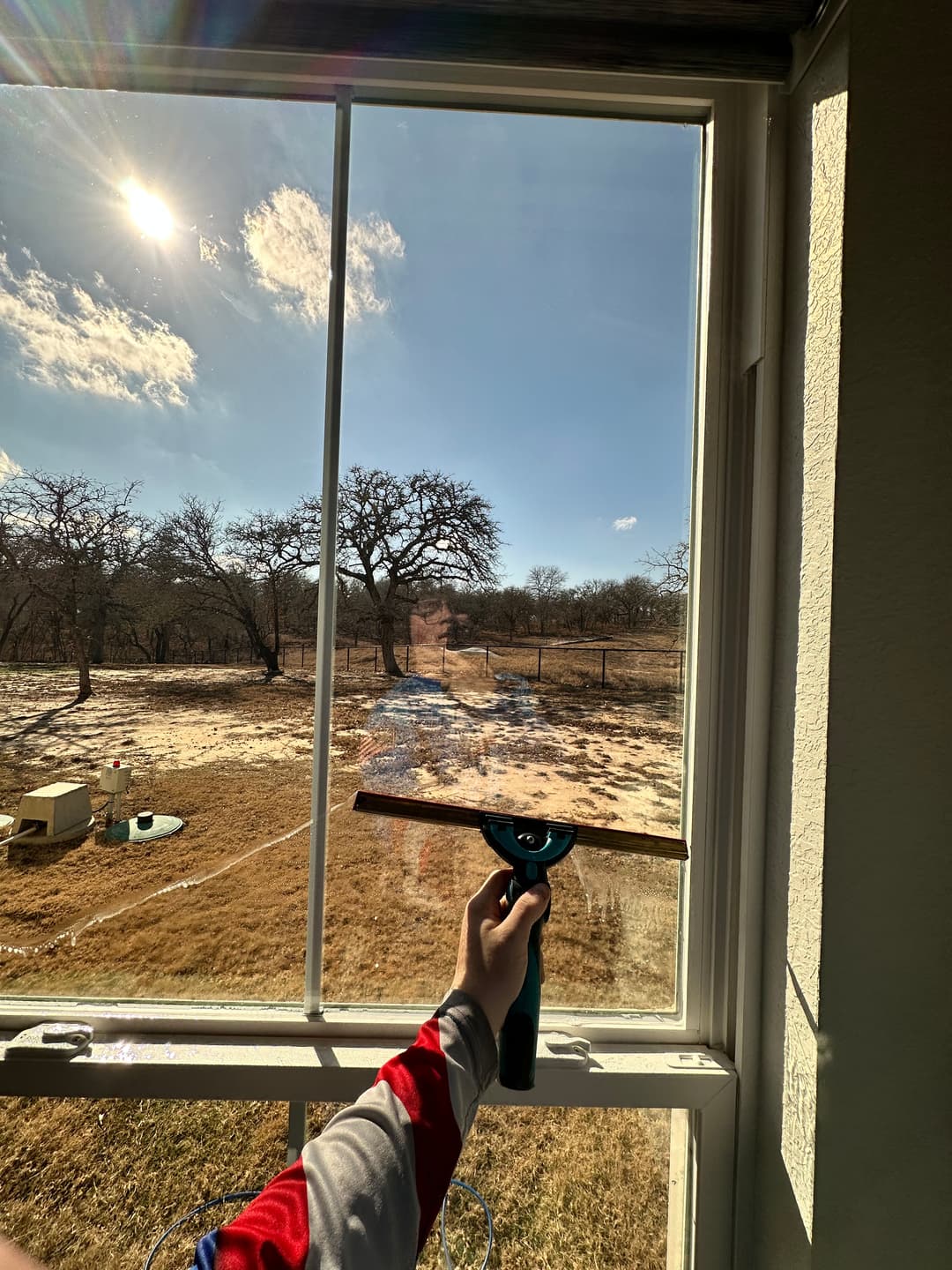 Person cleaning a window with a squeegee, with a sunny landscape visible outside.
