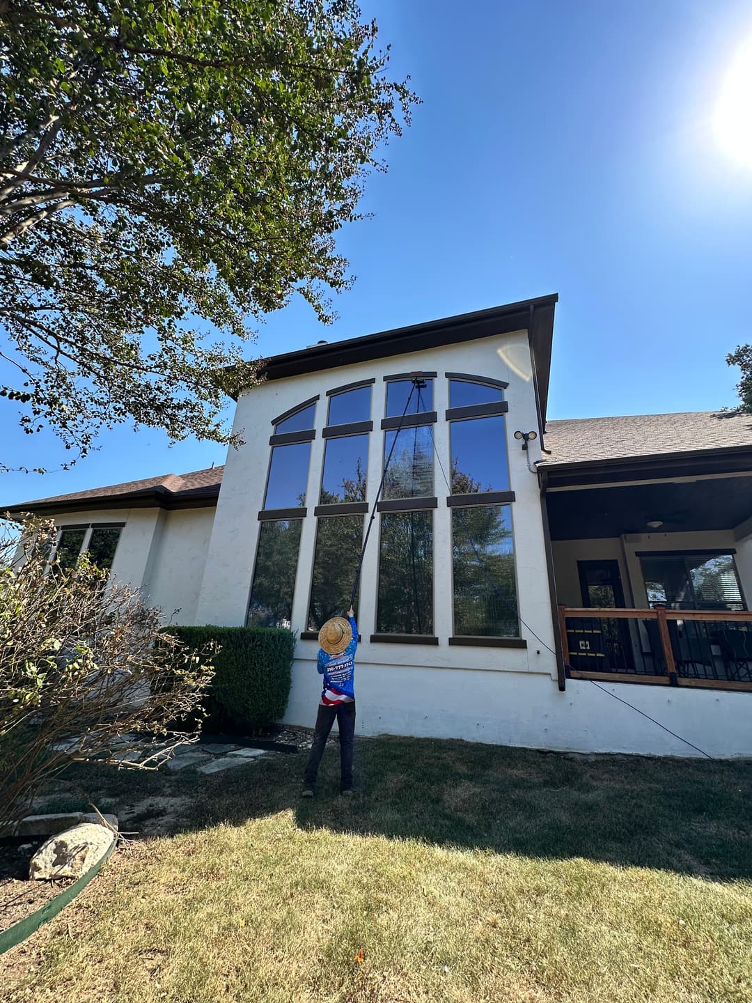 Person cleaning large windows on a house with a telescopic pole under clear blue sky.