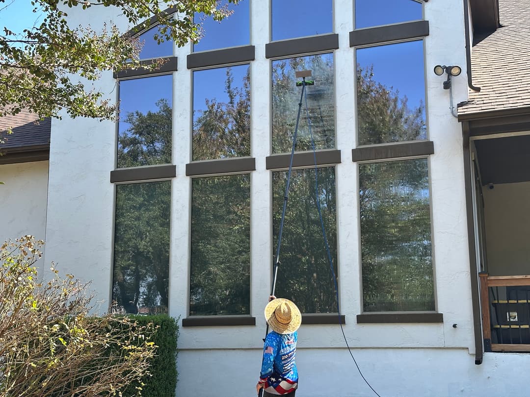 Person cleaning large windows on a home with a pole-mounted squeegee on a sunny day.