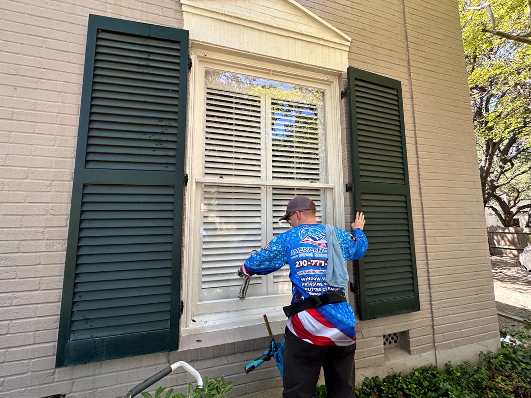 Window cleaning service professional using equipment on a house exterior with green shutters.