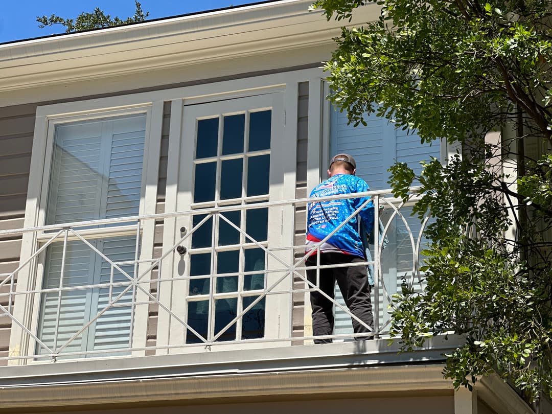 Person in blue shirt working on balcony of a house with trees in the foreground.