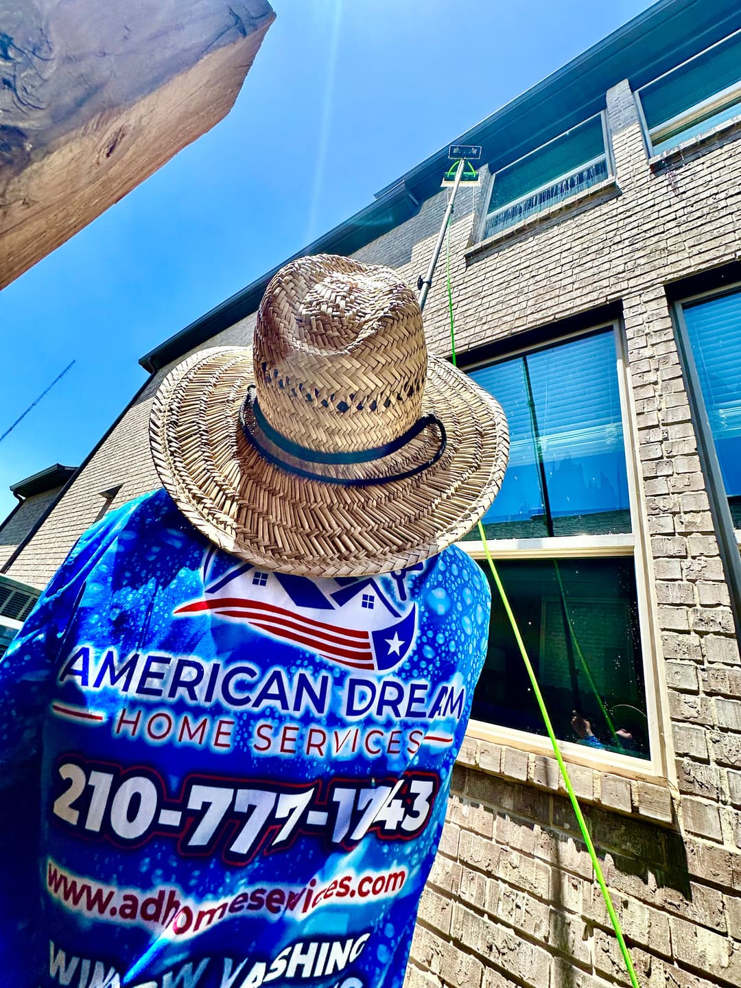 Window cleaning service by American Dream Home Services, worker in straw hat using equipment.