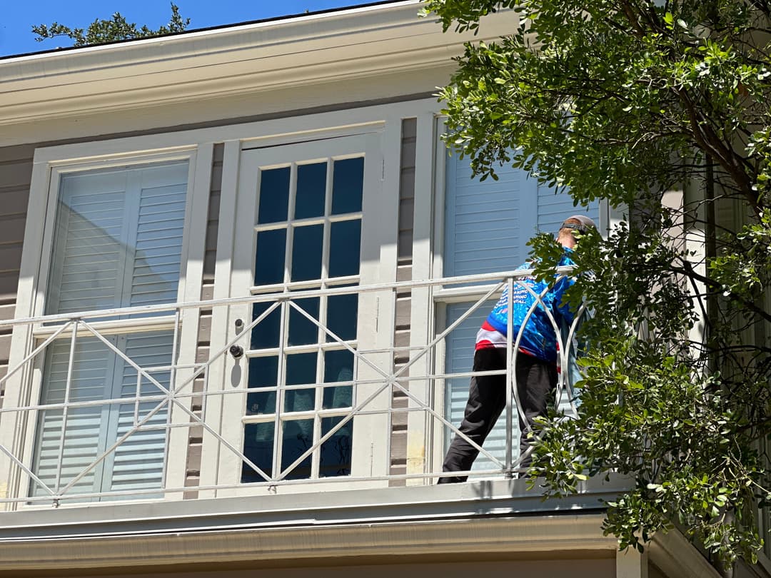 Person cleaning balcony of a house with colorful jacket, surrounded by greenery.