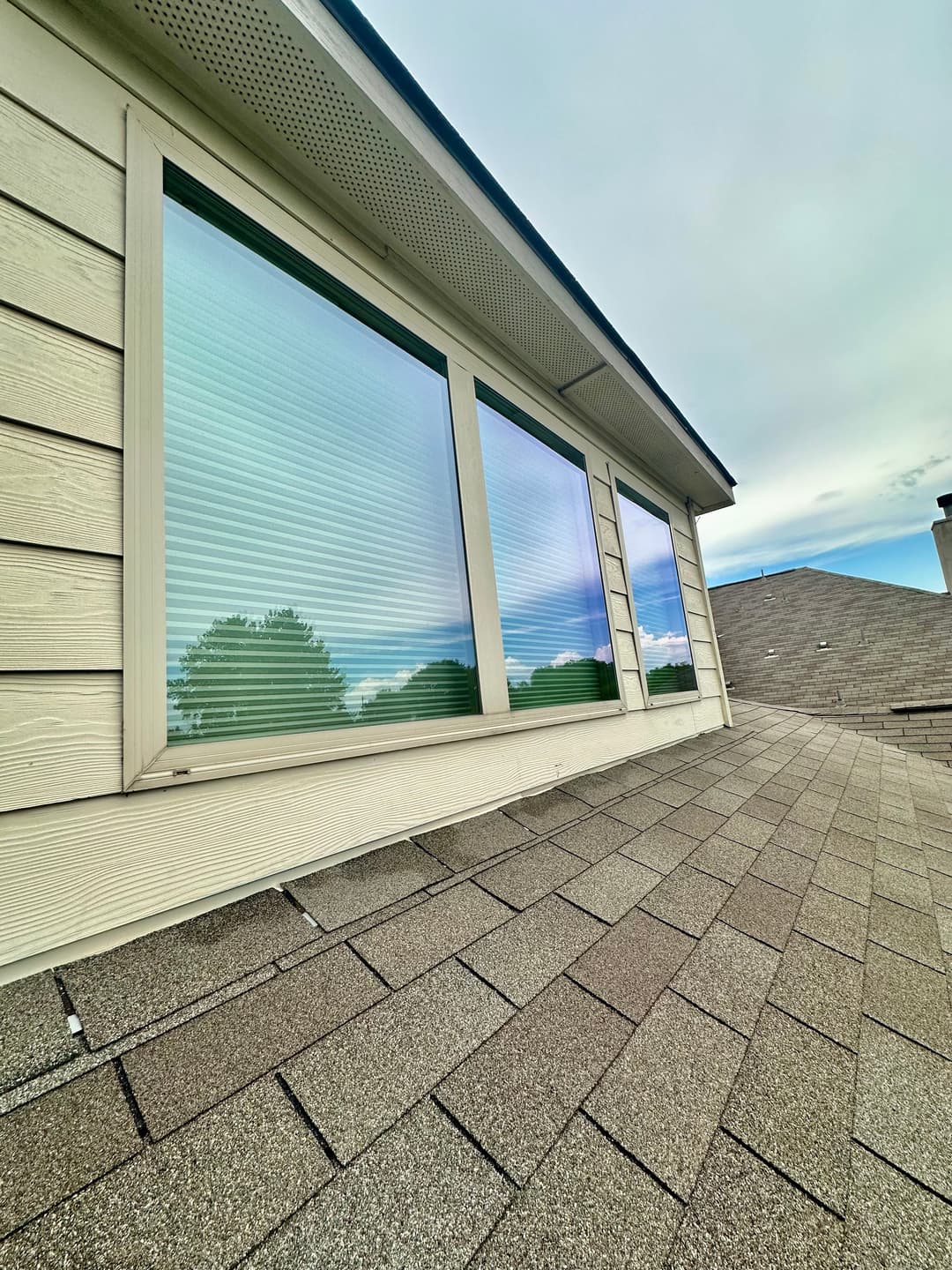 Windows with blinds reflecting the sky on a sloped roof with shingles.
