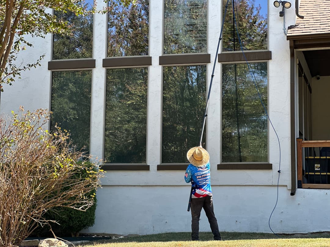 Person cleaning large windows on a house using a long pole and wearing a straw hat.