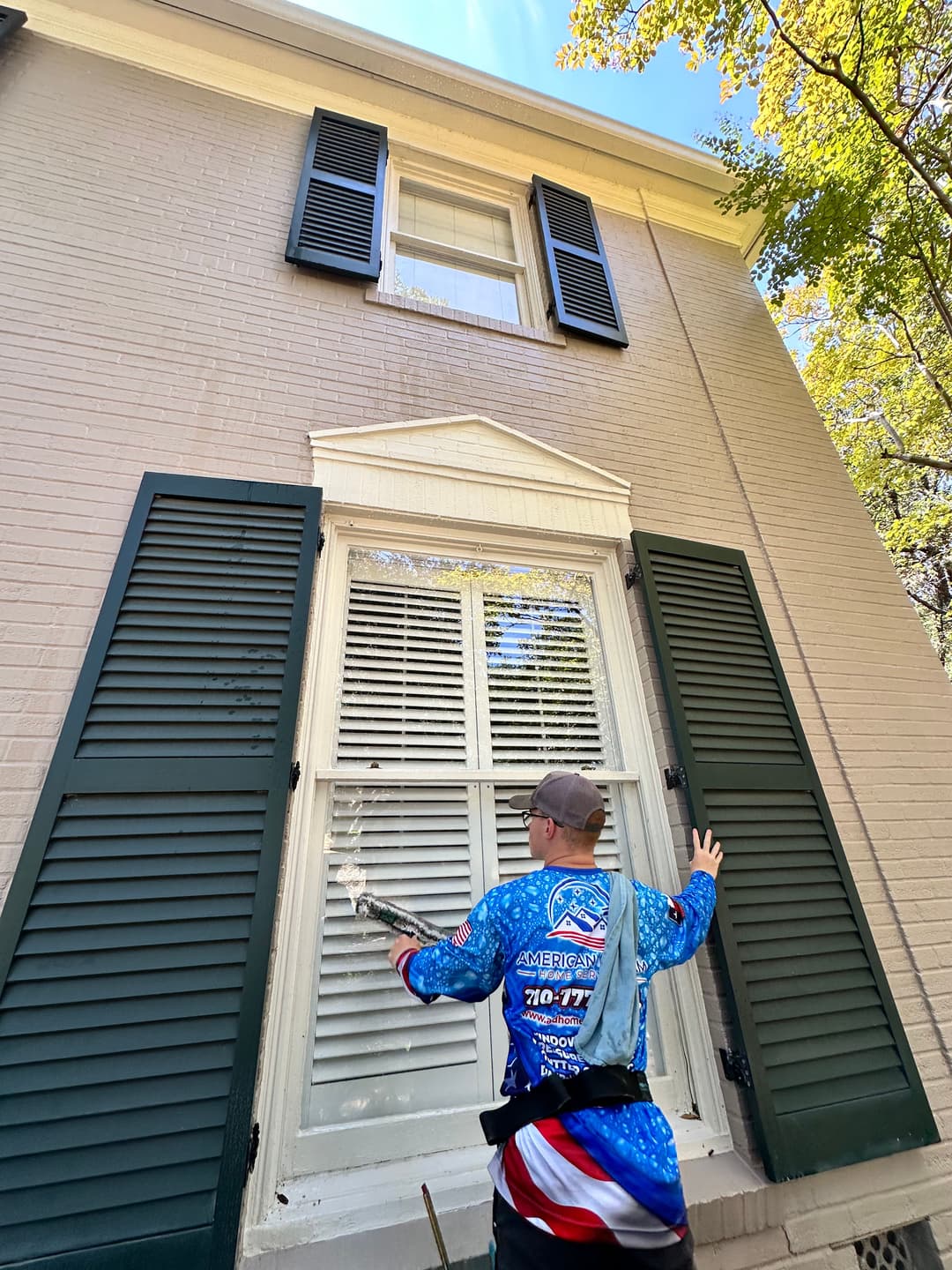 Person cleaning exterior window of a house with green shutters on a sunny day.