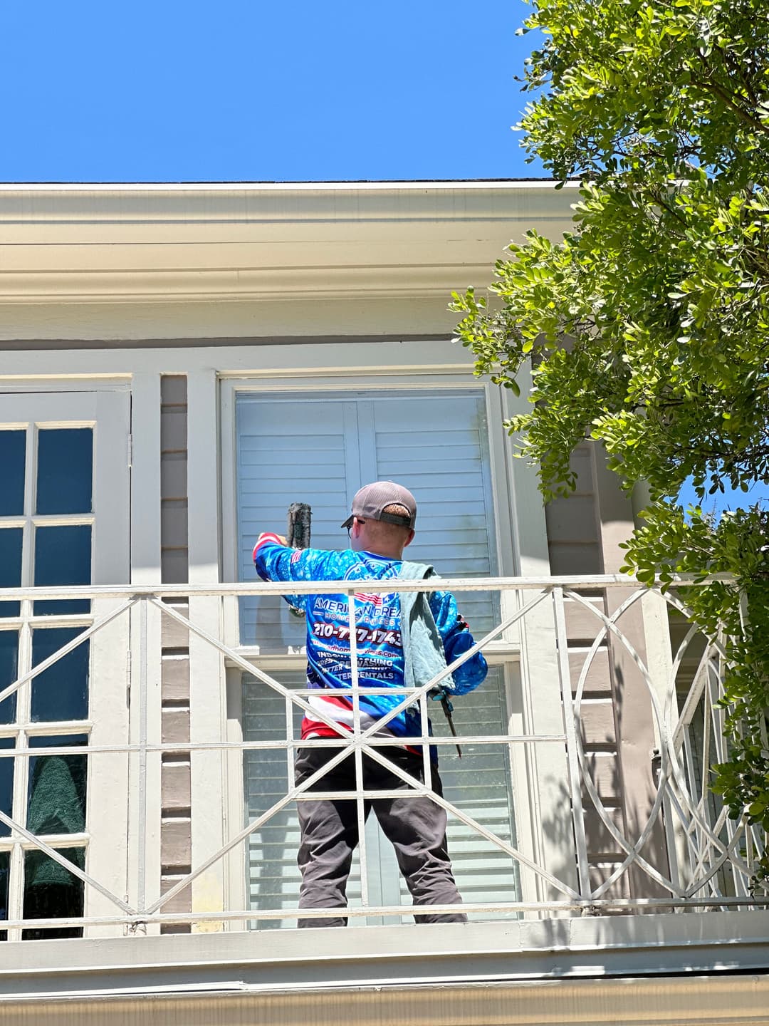 Worker cleaning windows on a balcony under a clear blue sky, wearing a colorful shirt.