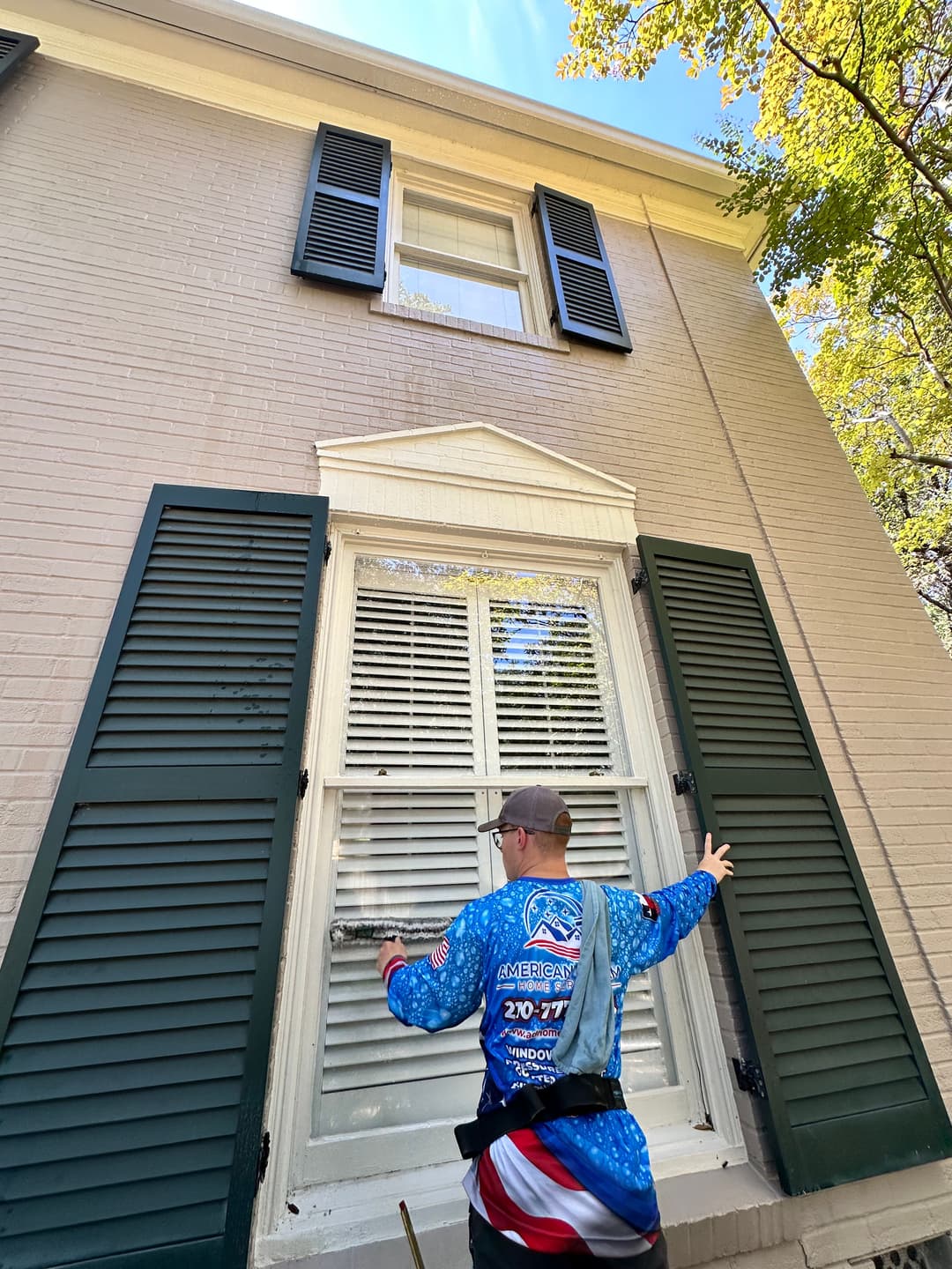 Window cleaning professional in colorful shirt working on second-story window of home.