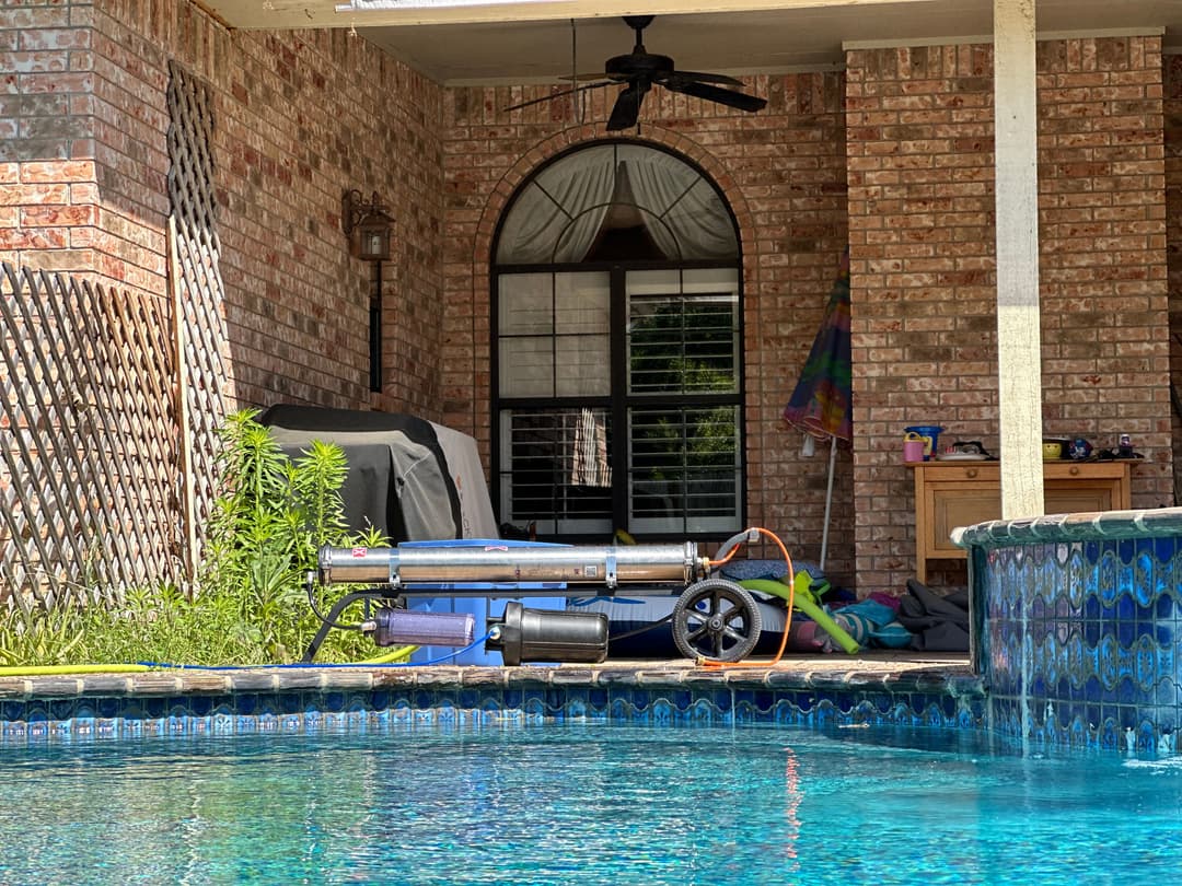 Pool cleaning equipment by a tiled pool with a brick patio and shaded area in the background.