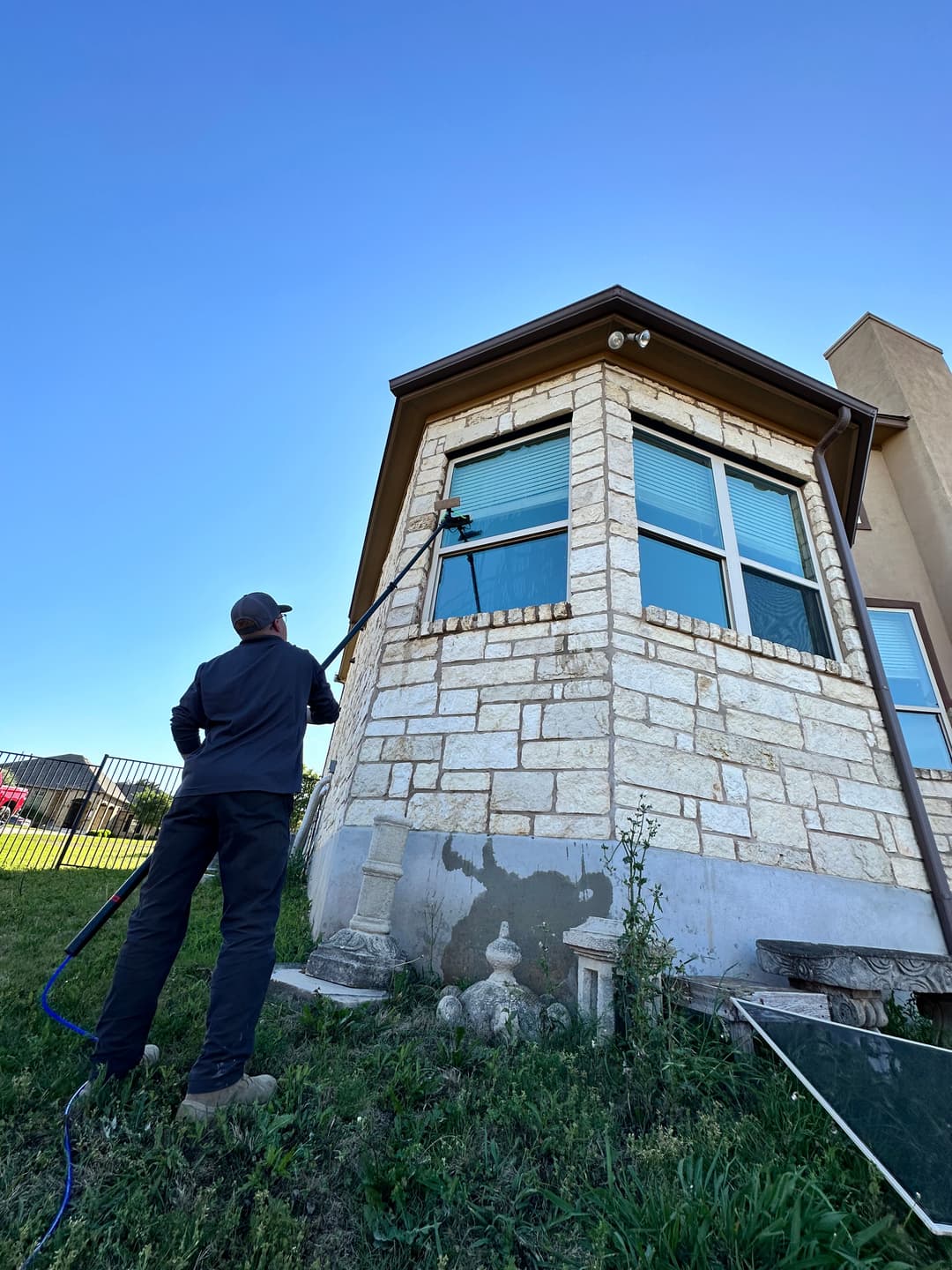 Person using a pole to clean windows on a stone house under a clear blue sky.