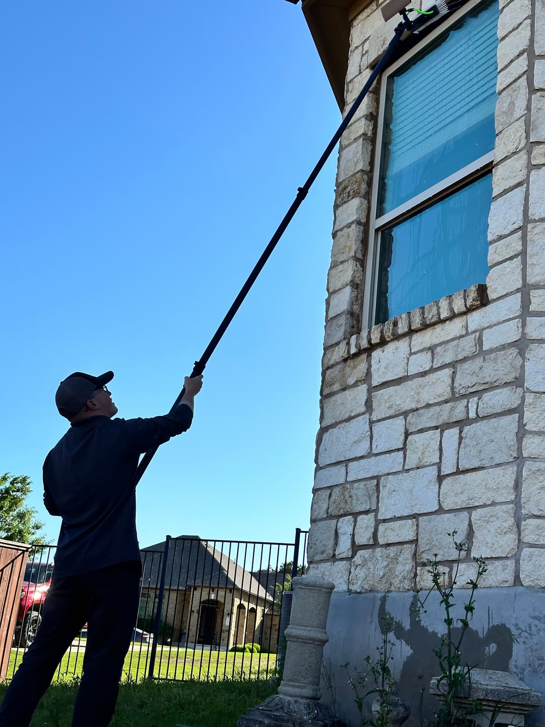 Person cleaning a second-story window with a long pole in bright sunlight.