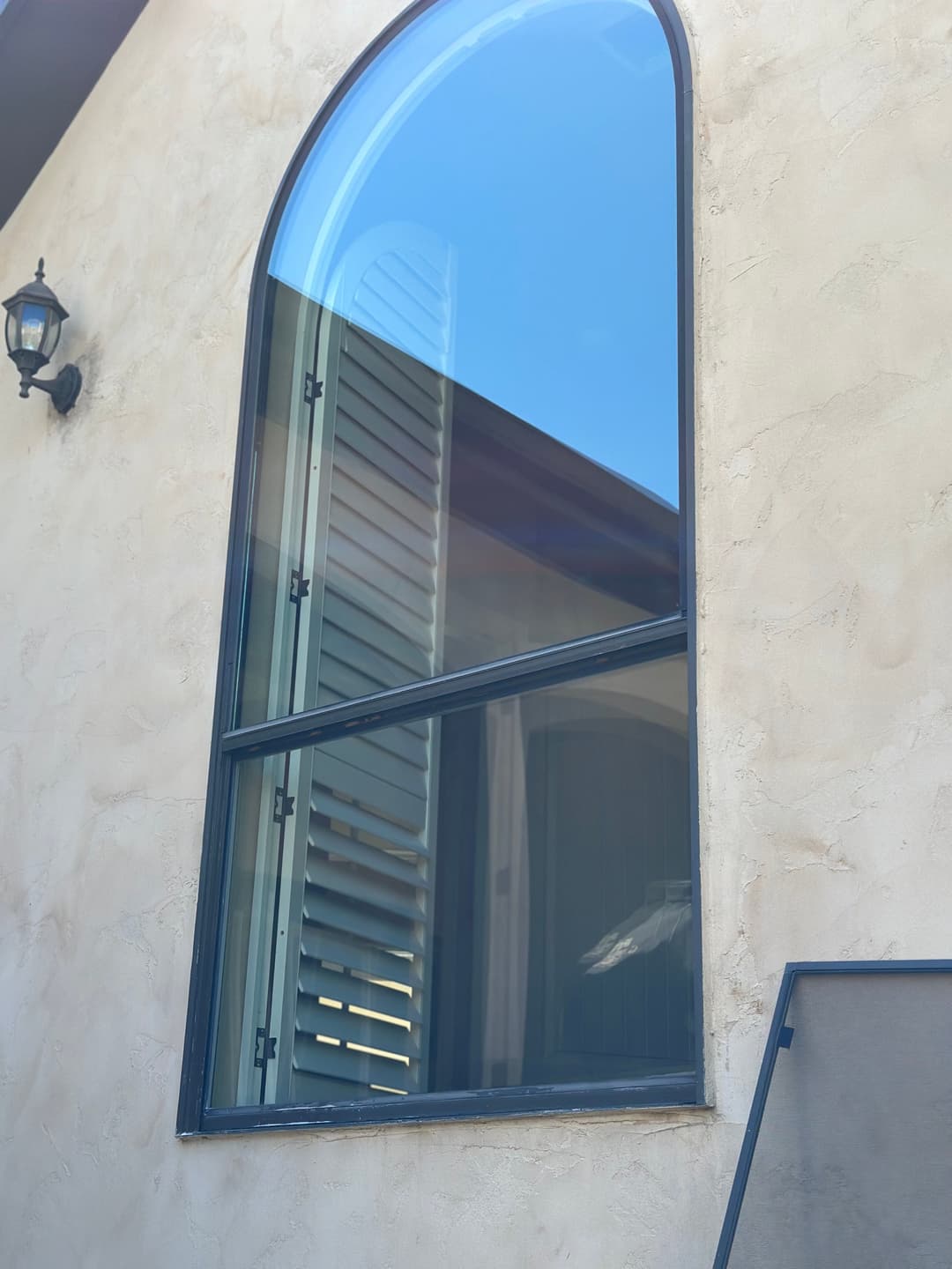 Large arched window with dark frame, featuring wooden shutters and blue sky reflection.