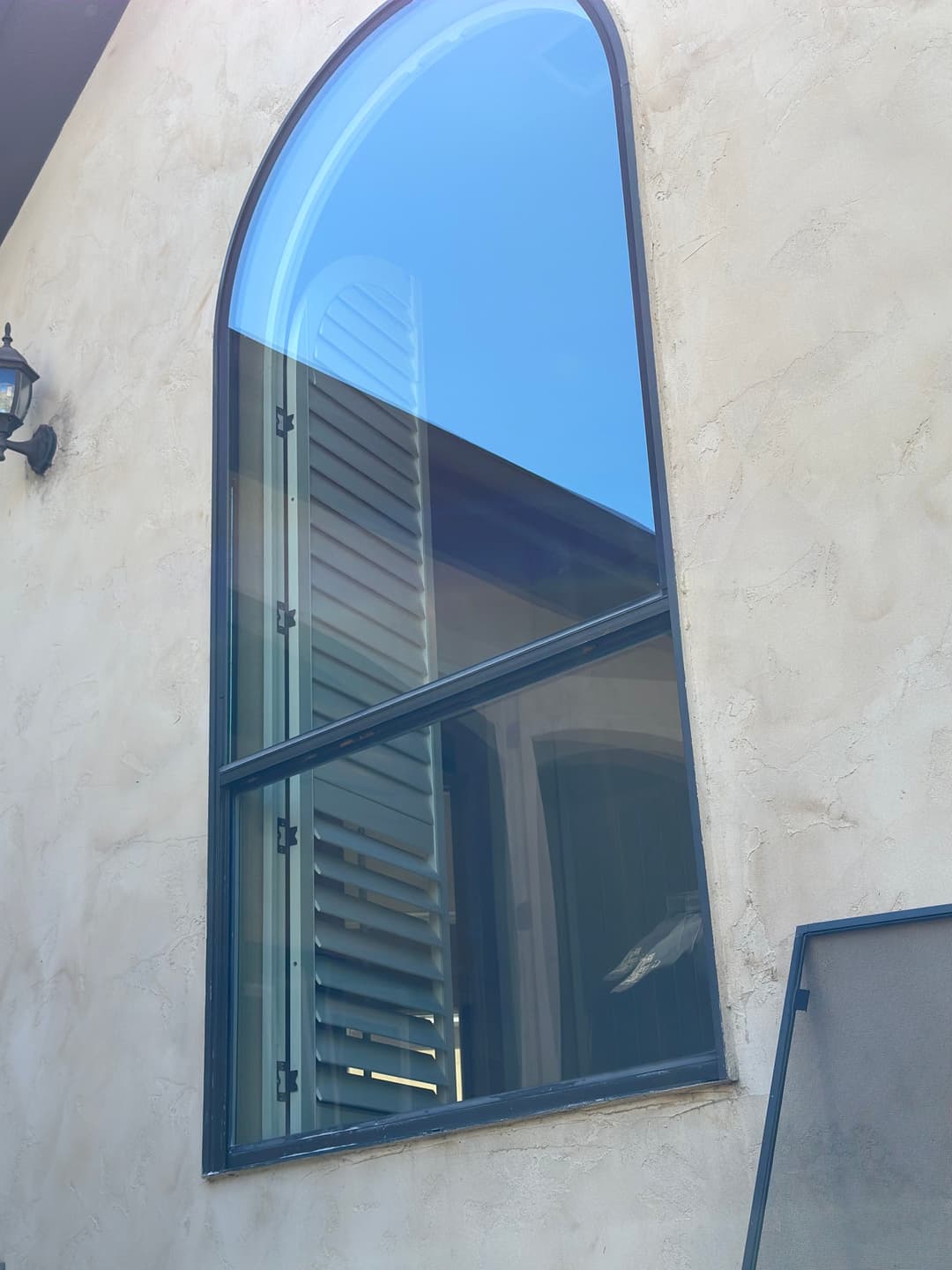 Modern arched window with reflective glass on a textured beige wall. Shutters partially open.