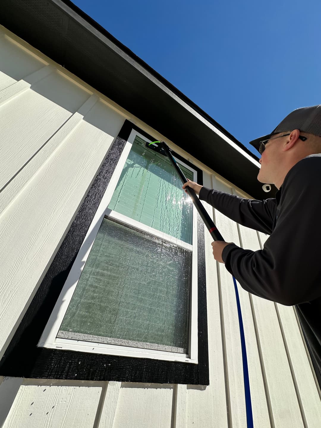 Person cleaning window of a house with a long squeegee under blue sky.