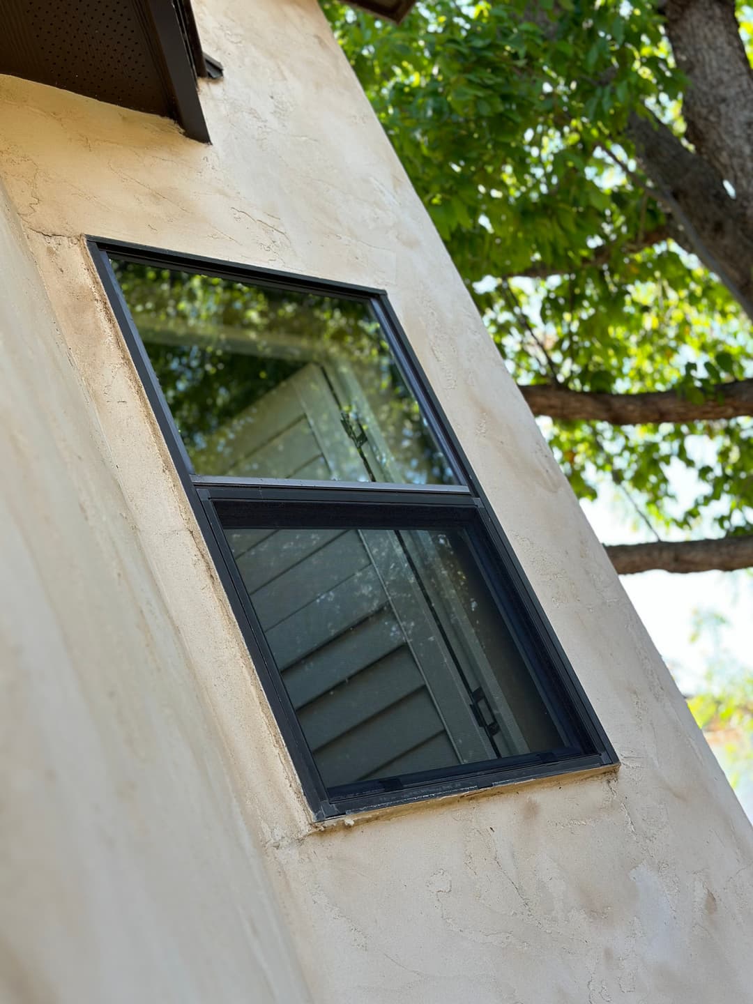 Black-framed window reflecting trees on a textured beige exterior wall.