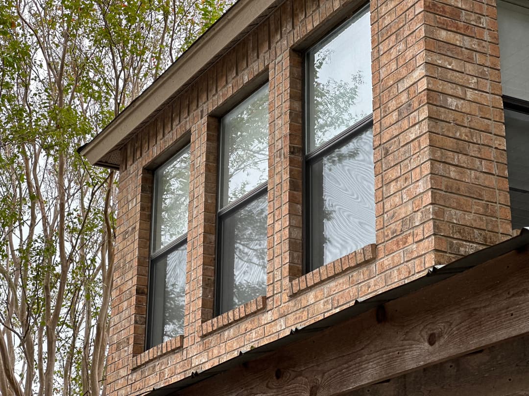 Modern brick house with three large windows and greenery in the background.