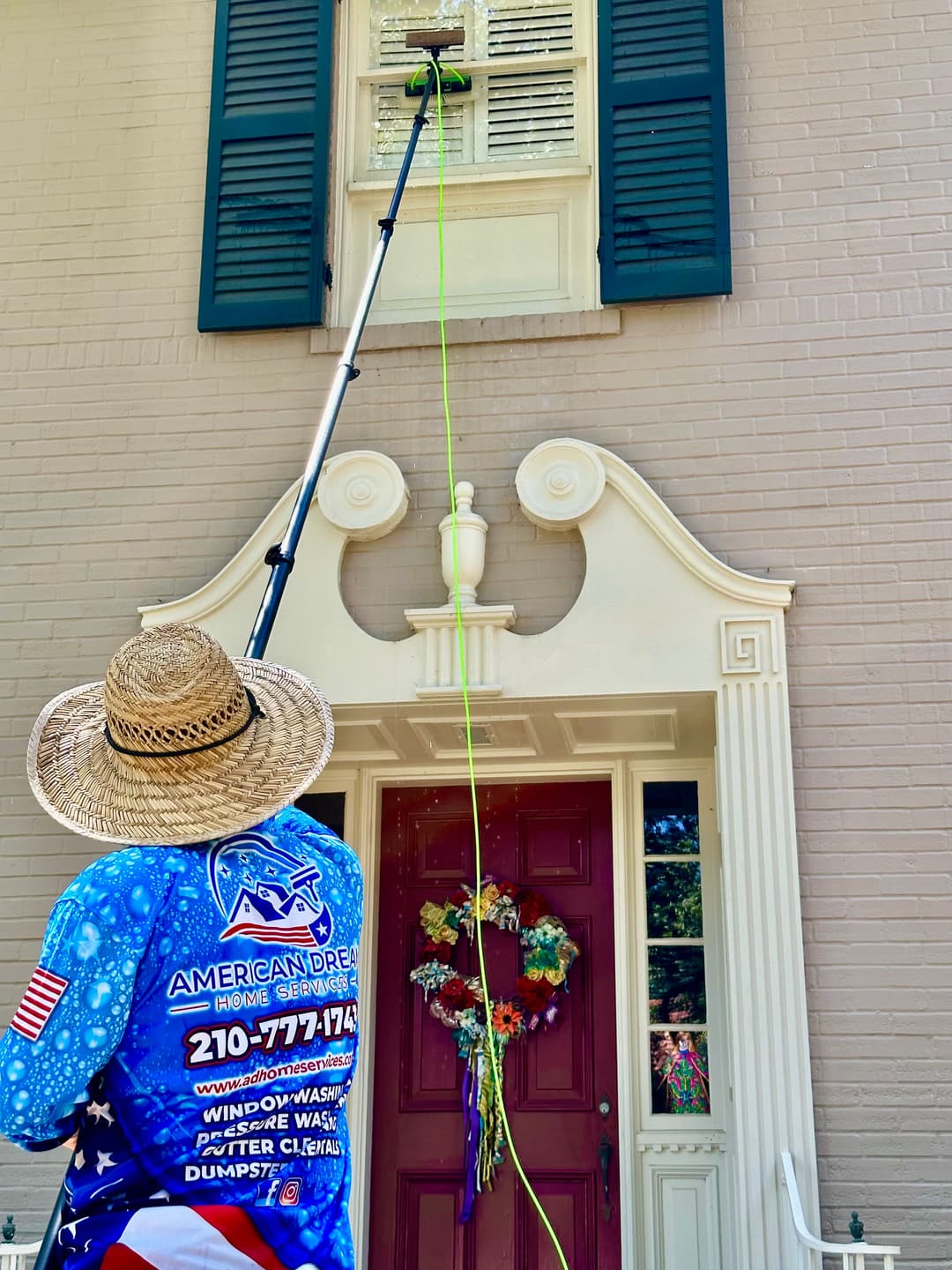 Man in a straw hat uses a pole to clean window shutters on a brick home.