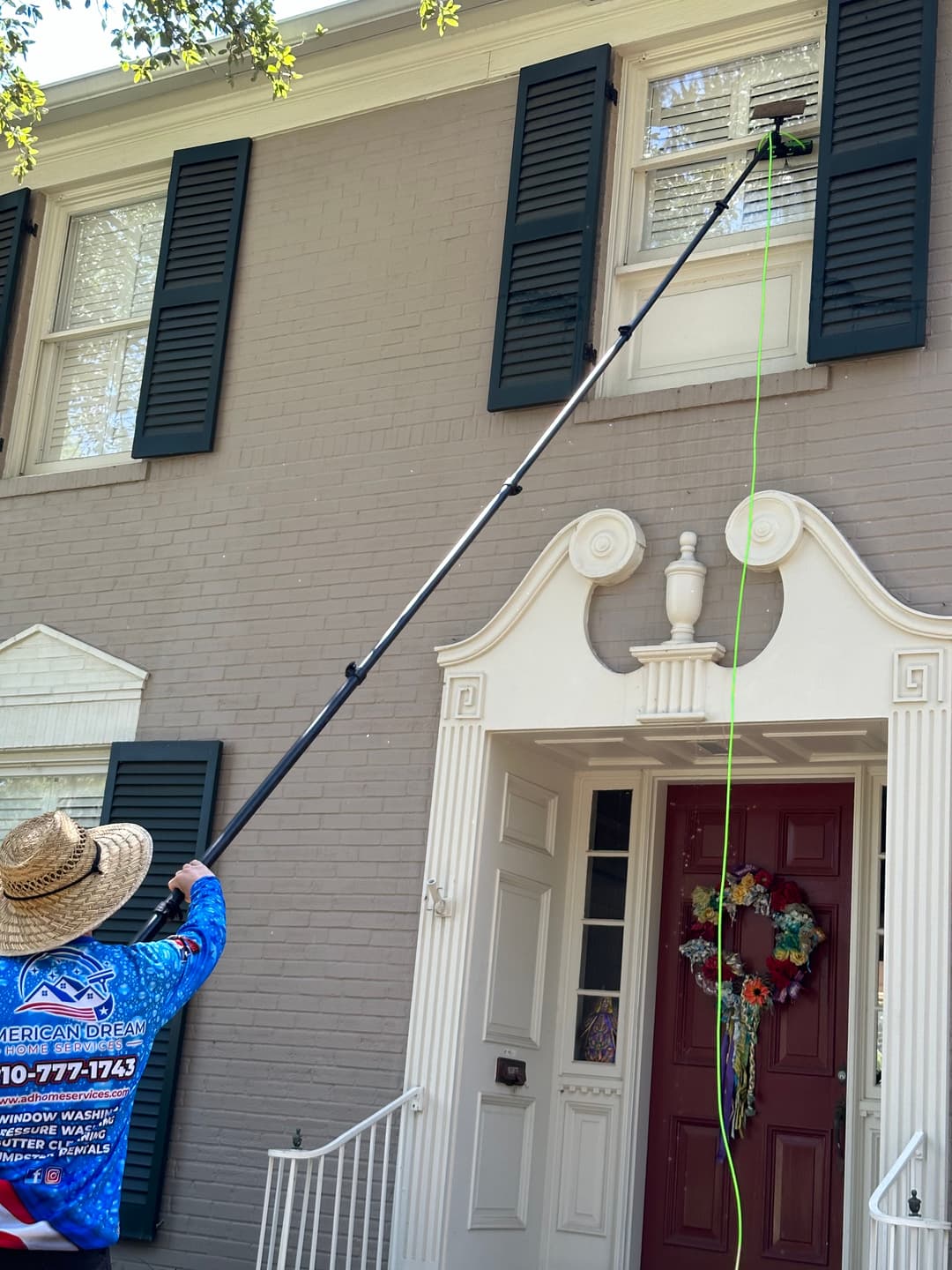 Worker using a long pole to clean a window on a gray brick house with white trim.