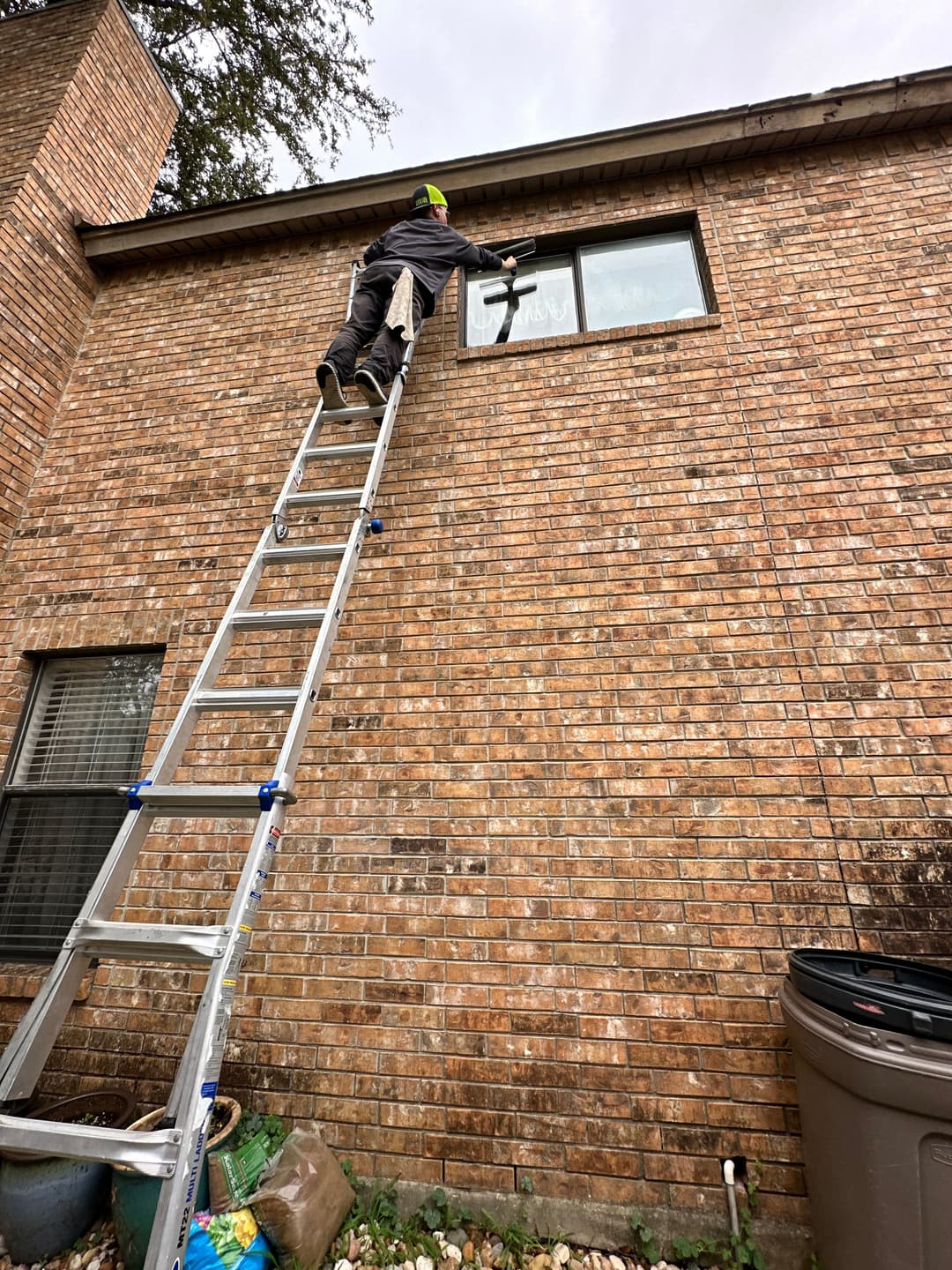 Person using a ladder to clean a window on a brick building exterior.