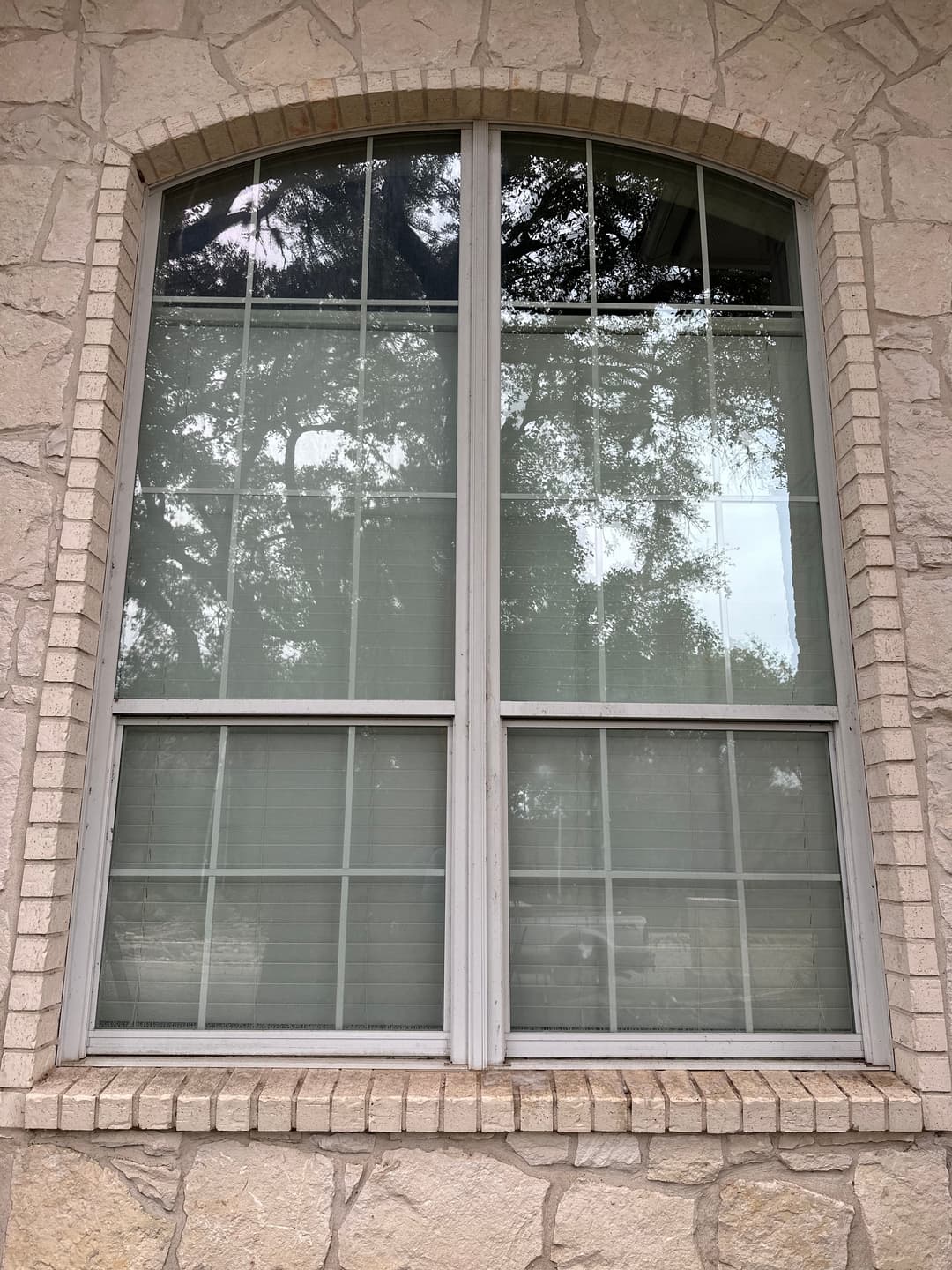 Two-pane window with grilles, framed by stone wall, reflecting trees outside.