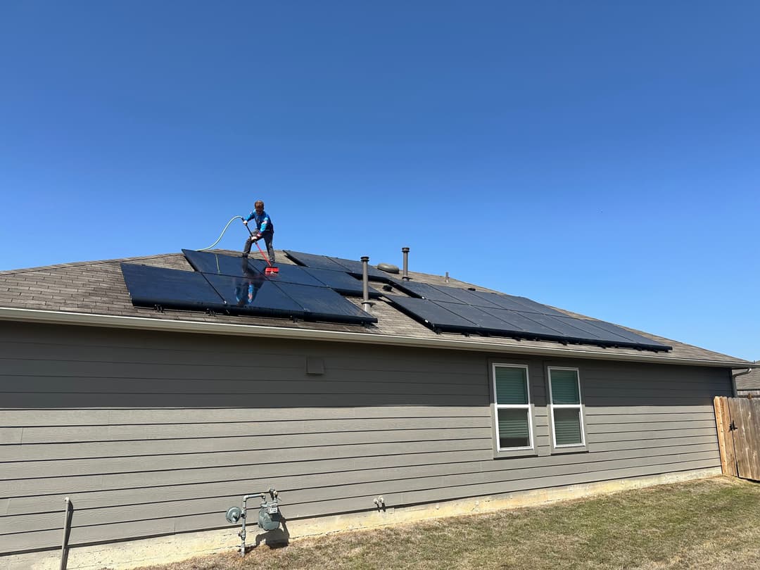 Worker cleaning solar panels on a house roof under a clear blue sky.