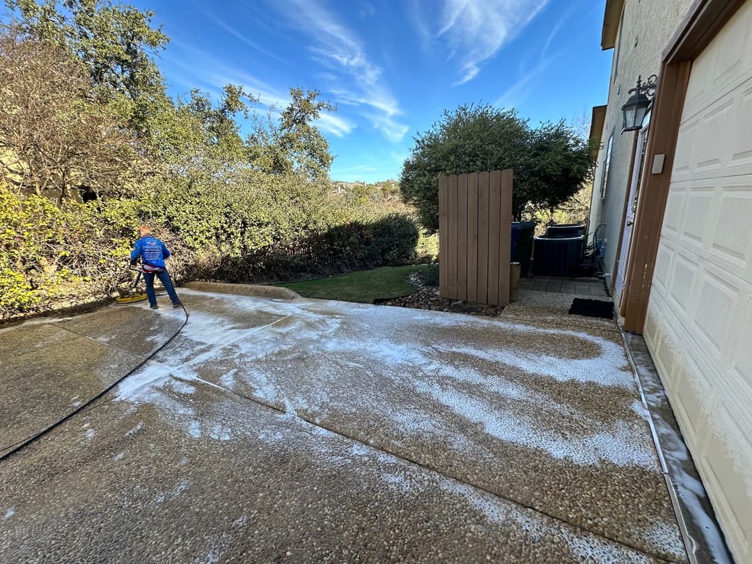 Person cleaning driveway with pressure washer, surrounded by greenery and blue sky.