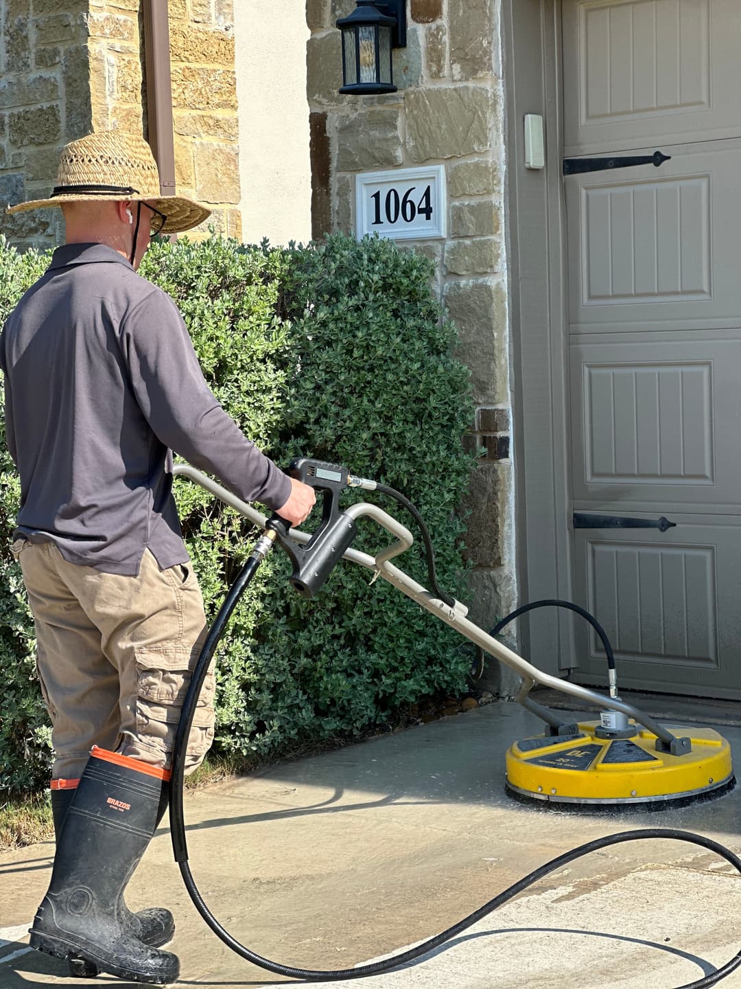 Person in hat using a surface cleaner for power washing a driveway outside a home.