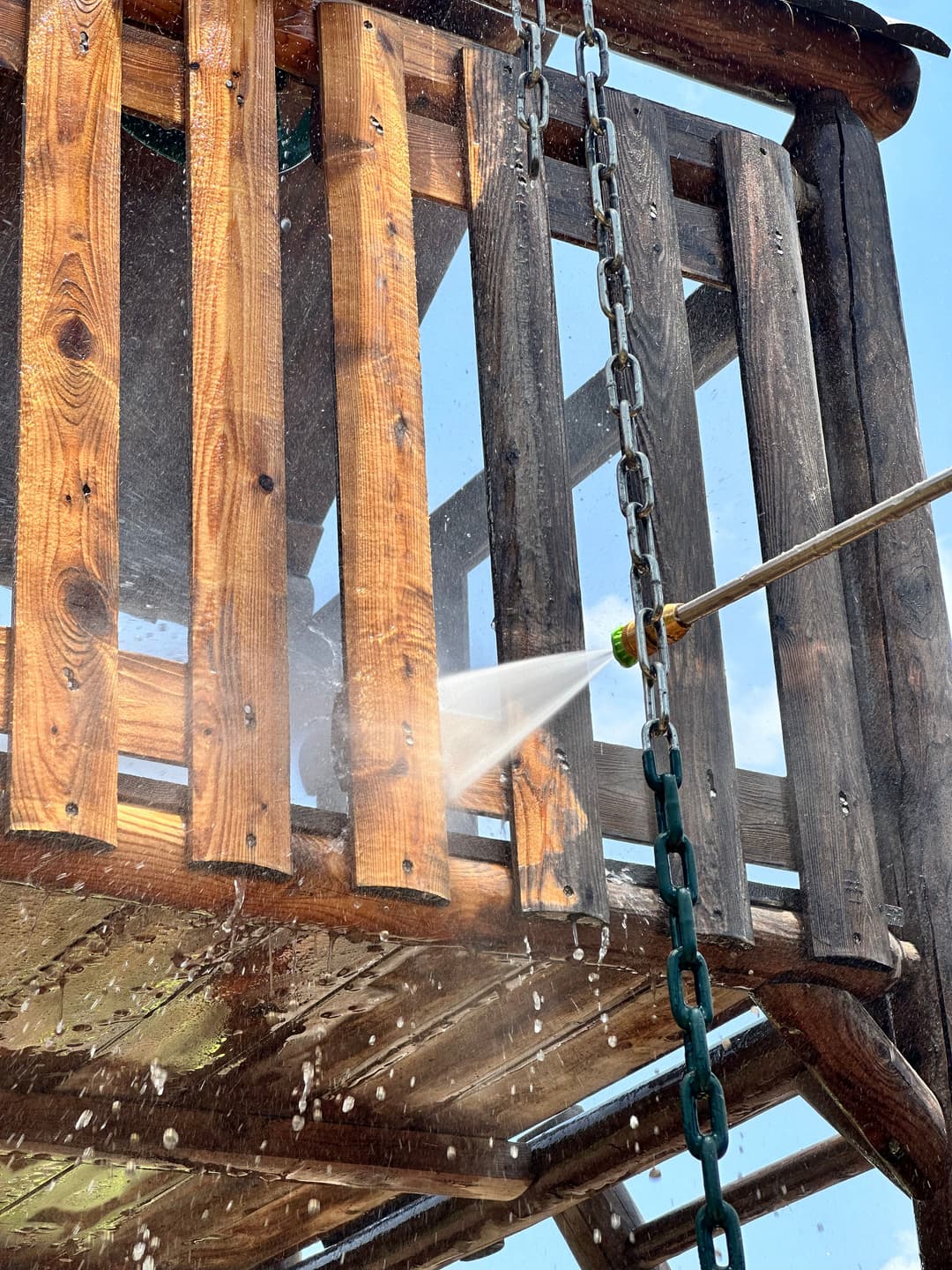 Water spraying from a hose onto a wooden playset, with sunlight shining from above.