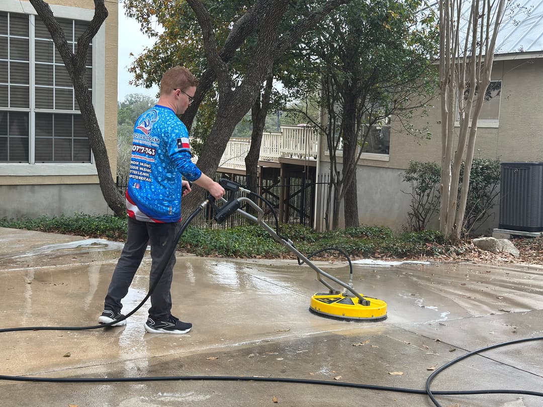 Person using a pressure washer on a driveway, wearing a blue shirt with a Texas flag design.