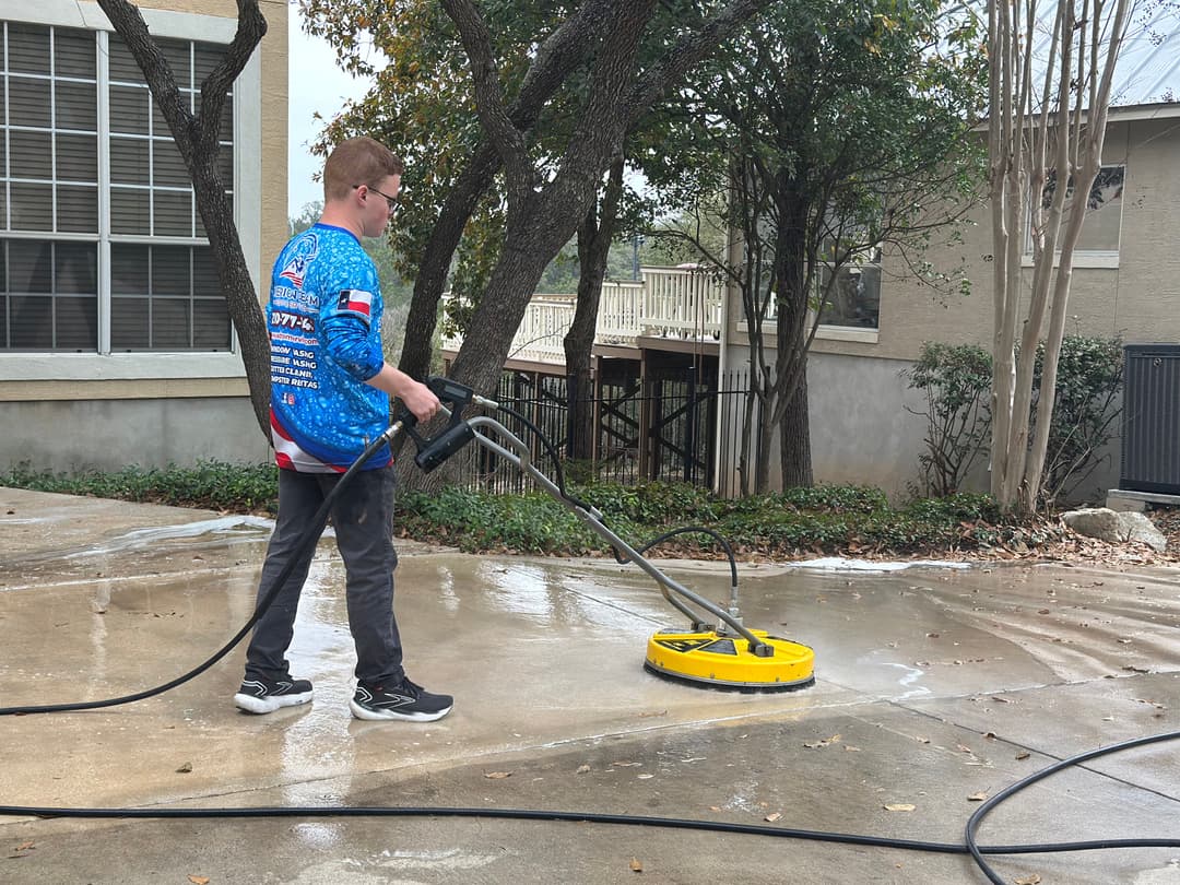 Man pressure washing a concrete patio with a yellow surface cleaner in a residential area.