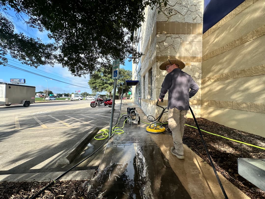 Worker using a pressure washer to clean a sidewalk beside a commercial building.