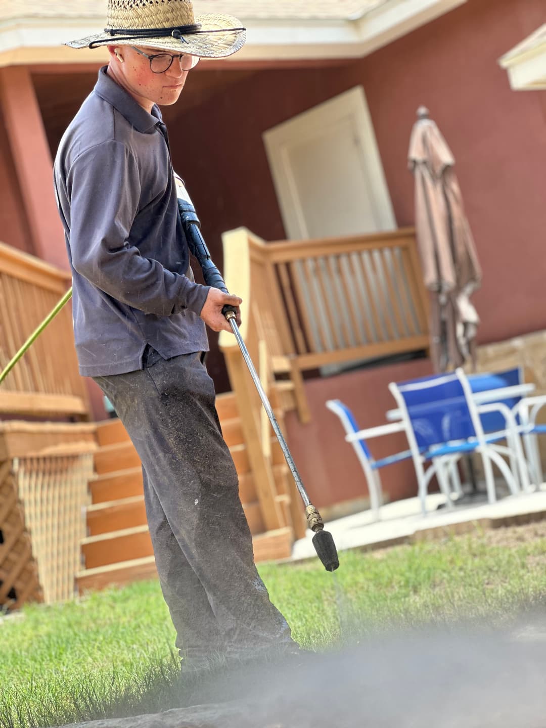Person in a straw hat using a pressure washer on a lawn near a house.