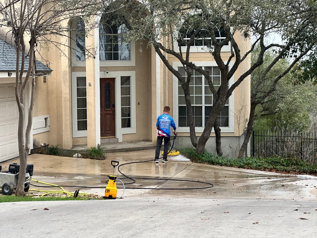 Person pressure washing a driveway in front of a two-story home surrounded by trees.