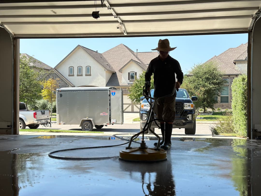 Person cleaning a garage floor with a machine, residential houses and trailer in background.