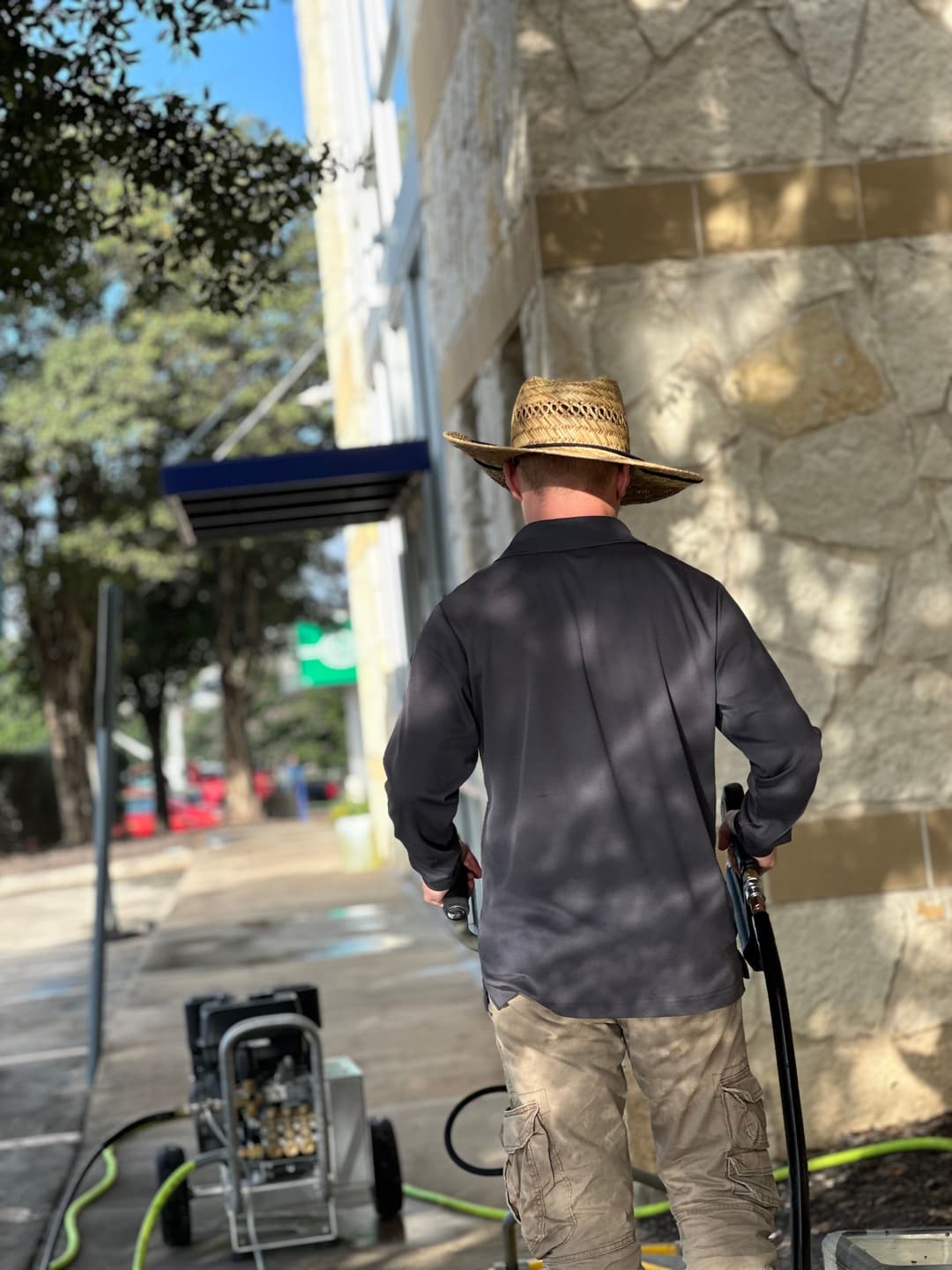 Man in a straw hat using a pressure washer on a stone building exterior.