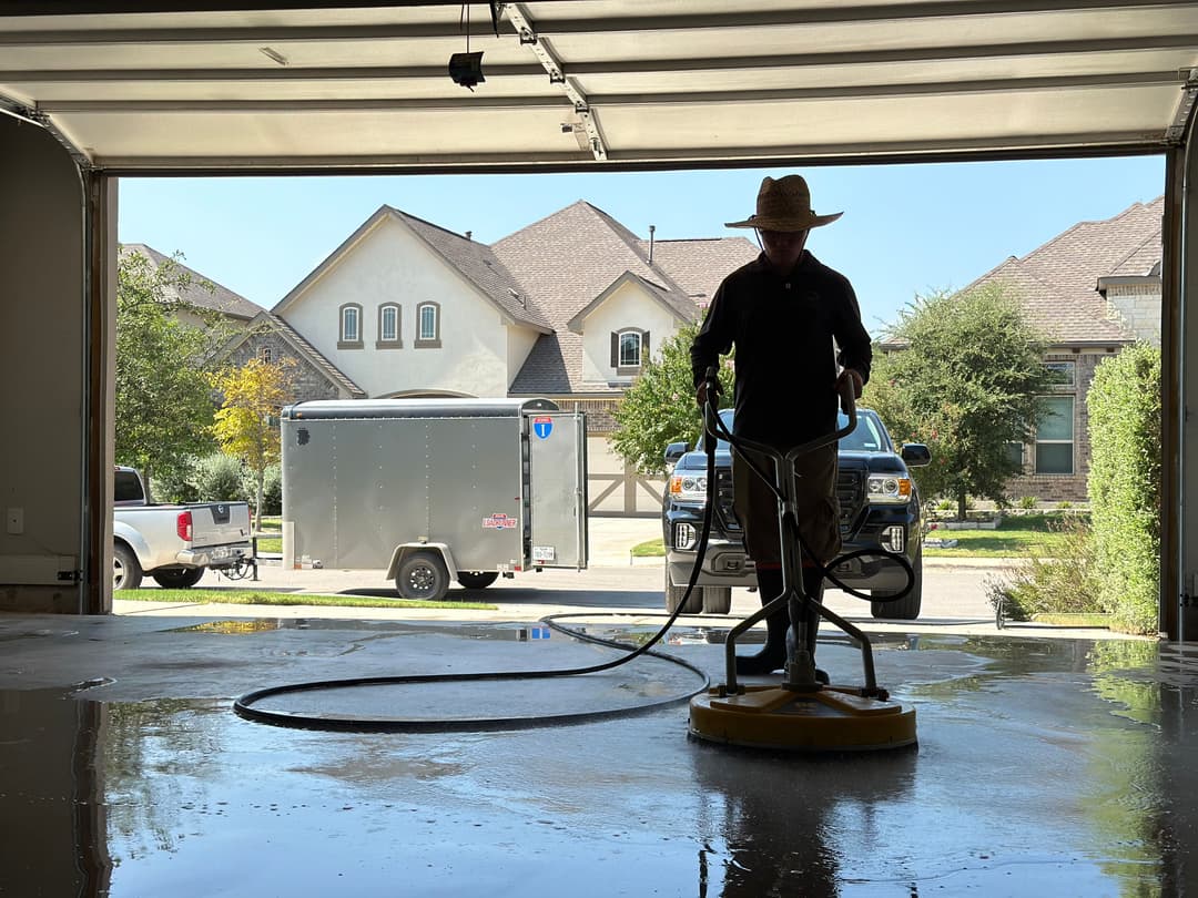 Person cleaning a garage floor with a floor buffer, houses and trucks visible in the background.