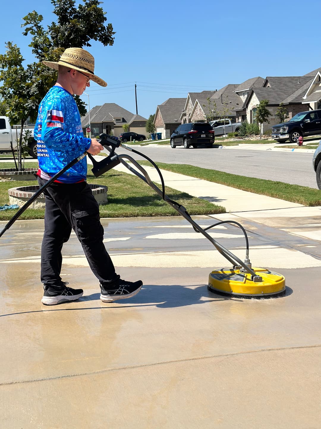 Person using a pressure washer on a driveway, wearing a straw hat and colorful shirt.