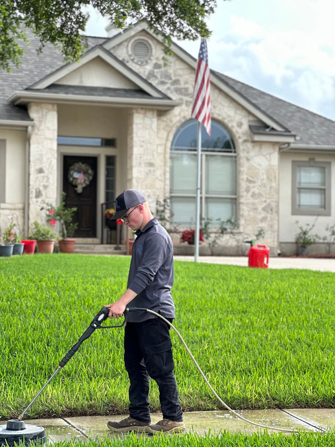 Man pressure washing a sidewalk in front of a stone house with an American flag.