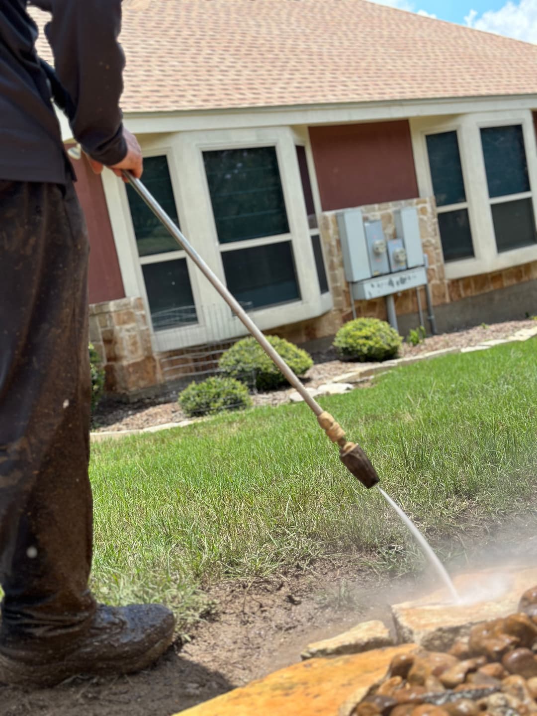 Person using a pressure washer on grass near a residential building with landscaping.