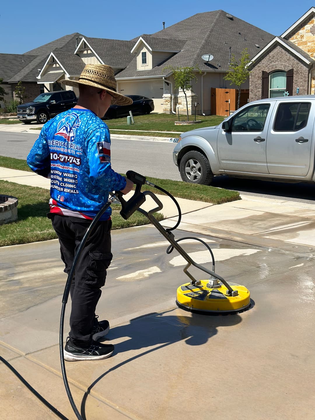 Man using a pressure washer on a driveway with a home and car in the background.