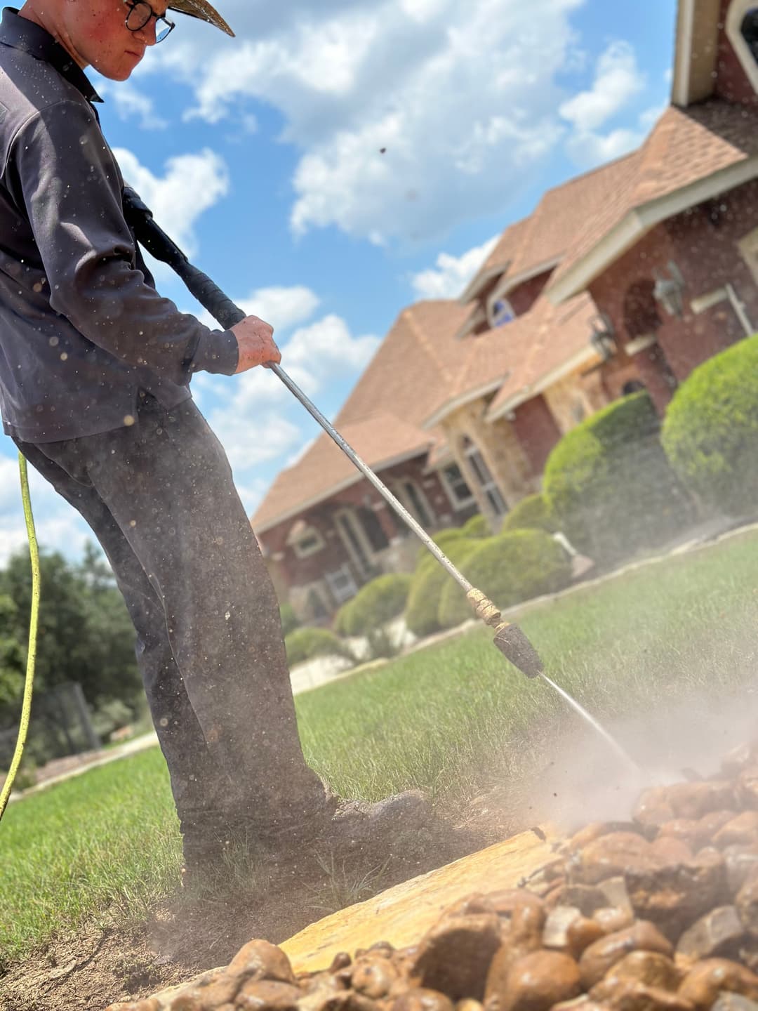 Person using a pressure washer on landscaping stones in a sunny yard with a house in background.