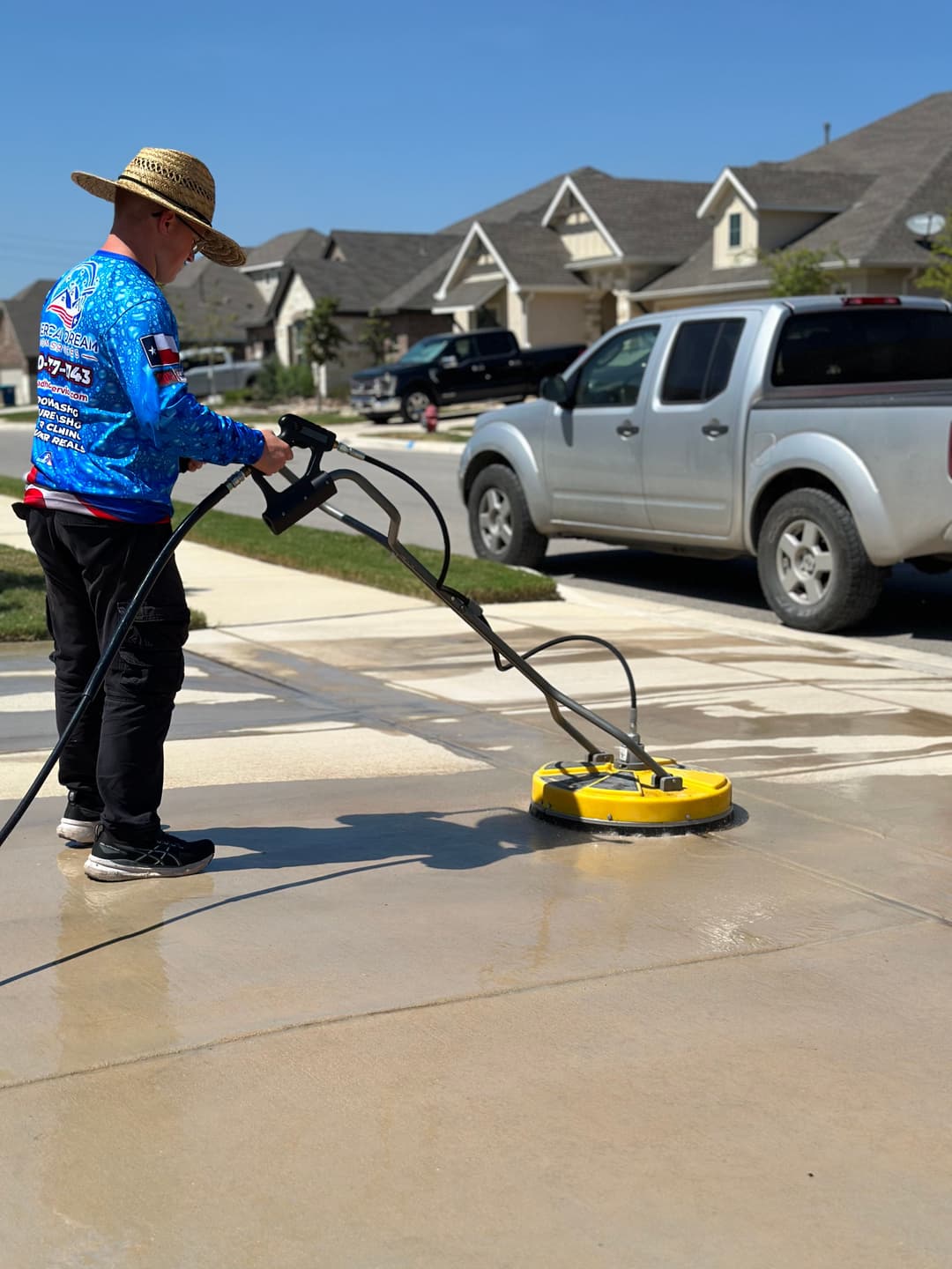 Man using a power washer to clean a driveway on a sunny day in a residential area.